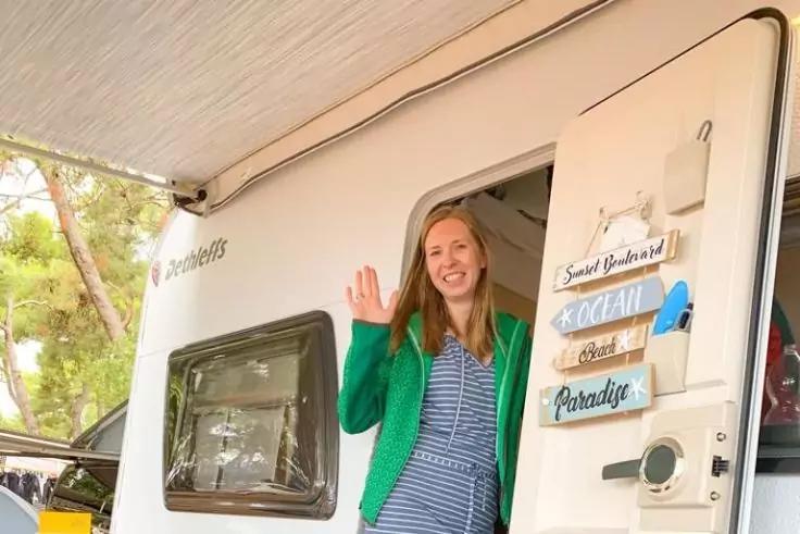 Person steht in ge&ouml;ffneter Wohnwagent&uuml;r, winkt, tr&auml;gt gr&uuml;nen Cardigan und gestreiftes Kleid; Wohnwagen mit Fenster und h&auml;ngenden Holzschildern an der T&uuml;r