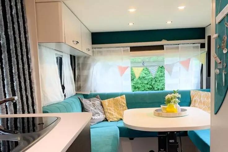 Innenraum eines Wohnwagens mit t&uuml;rkisfarbener U-Sitzgruppe um einen runden Tisch, wei&szlig;e Vorh&auml;nge am Fenster, Wimpelkette, gelbe Blumen in einer Schale auf dem Tisch, K&uuml;chenzeile links