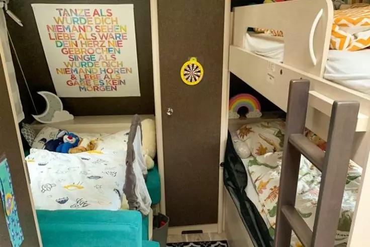 Kleiner Schlafraum mit zwei gegen&uuml;berliegenden Betten: links ein niedriges Bett mit t&uuml;rkisfarbener Seitenlehne, wei&szlig;er Bettw&auml;sche und blauem Stofftier; rechts ein Etagenbett mit Leiter, gemusterter Bettw&auml;sche und Regenbogenkissen; an der linken Wand