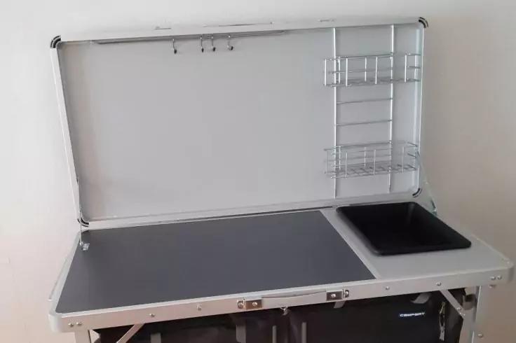 Offener Metallkoffer als Tisch; obere Klappe ge&ouml;ffnet mit drei Haken und zwei Drahtregalen rechts; graue Arbeitsfl&auml;che mit schwarzer, rechteckiger Einlasswanne rechts; Metallgestell und schwarze Tasche darunter; wei&szlig;er Hintergrund