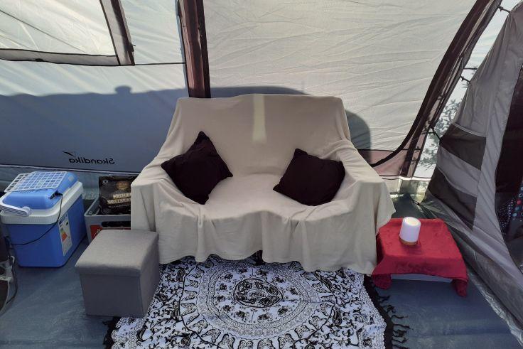 Sofa mit wei&szlig;er Decke und zwei dunklen Kissen in einem Zelt; links eine blaue K&uuml;hlbox, rechts ein kleiner roter Tisch mit wei&szlig;er Tasse; gemusterter Teppich davor