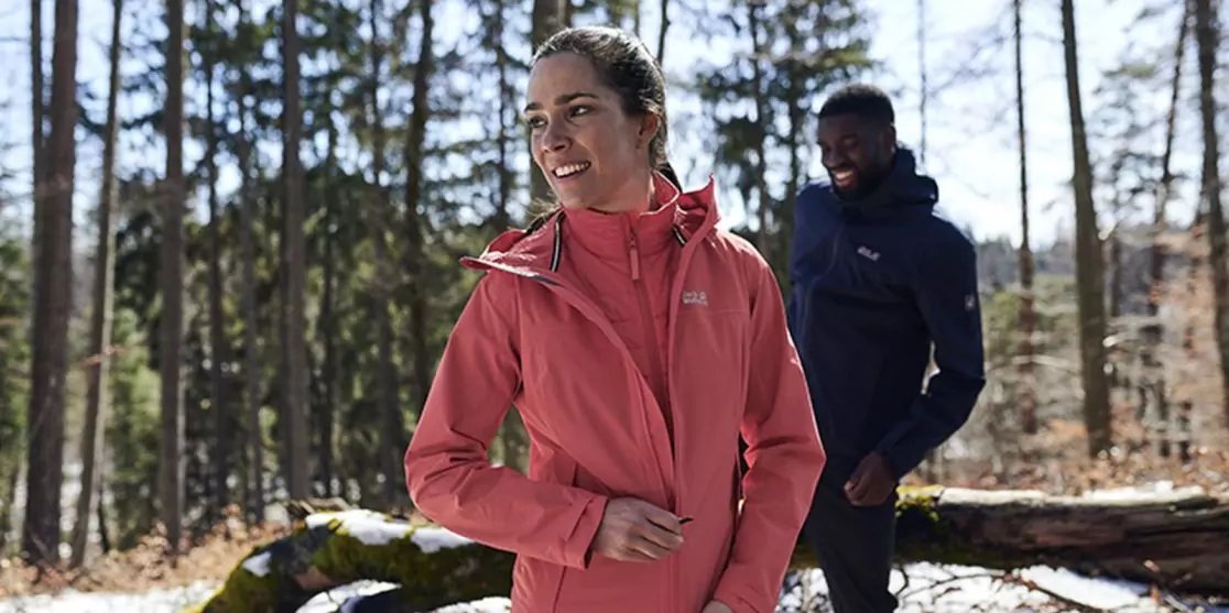 Frau in rosa Outdoorjacke l&auml;chelnd im Vordergrund, Mann in dunkler Jacke im Hintergrund, beide auf einem Waldweg mit B&auml;umen und Schneeresten