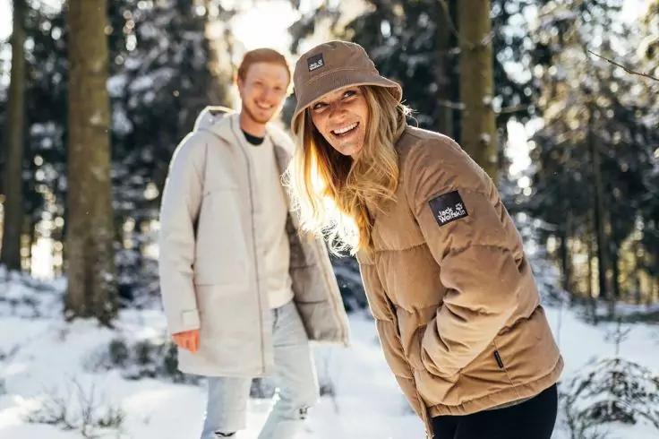 Zwei Personen in Winterkleidung in einem verschneiten Wald, eine trägt eine braune Jacke und einen Anglerhut, die andere eine hellfarbene Jacke