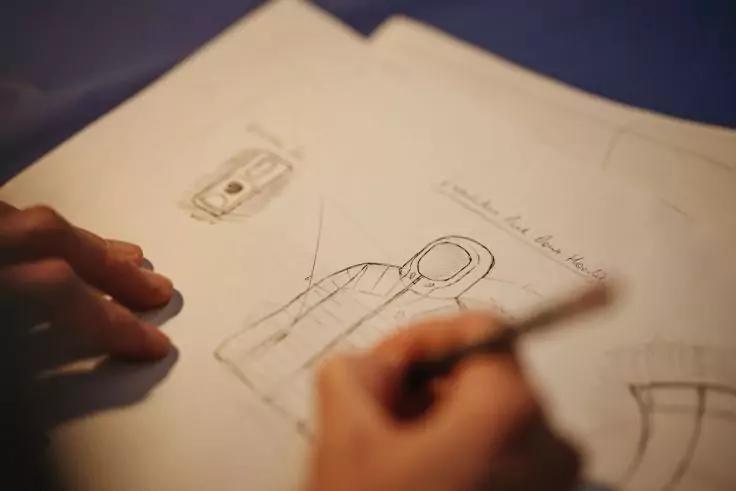 Hände, eine hält einen Bleistift und zeichnet auf Papier Skizzen eines Kleidungsstücks mit Kapuze; unscharfer dunkelblauer Hintergrund