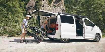 Wei&szlig;er Kleinbus auf Schotter, Heckklappe und Schiebet&uuml;r offen; eine Person mit Hut schiebt ein Fahrrad in den Laderaum, eine weitere Person sitzt im ge&ouml;ffneten Heck; B&auml;ume und Felswand im Hintergrund