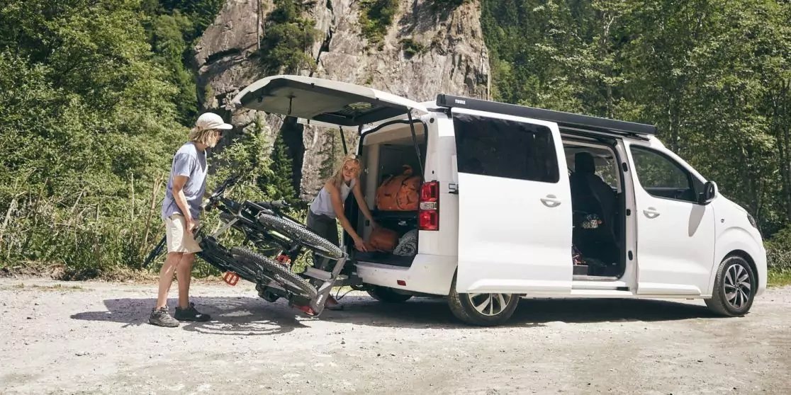 Wei&szlig;er Kleinbus auf Schotter, Heckklappe und Schiebet&uuml;r offen; eine Person mit Hut schiebt ein Fahrrad in den Laderaum, eine weitere Person sitzt im ge&ouml;ffneten Heck; B&auml;ume und Felswand im Hintergrund
