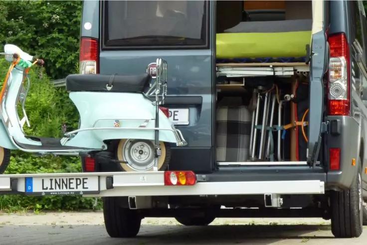Offenes Heck eines Transporters mit einer hellblauen Retro-Roller auf einer Ladeplattform, zusammengeklappte St&uuml;hle und gr&uuml;ner Polster im Innenraum