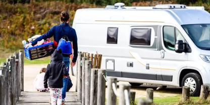 Erwachsene Person tr&auml;gt eine Kiste auf einem Holzsteg; zwei Kinder, eines mit blauer M&uuml;tze, gehen vor ihr; rechts steht ein wei&szlig;es Wohnmobil; Pfosten entlang des Stegs und D&uuml;nen im Hintergrund