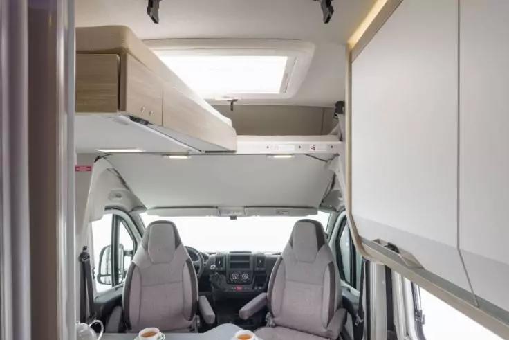 Innenansicht eines Wohnmobils mit zwei grauen Fahrersitzen, Lenkrad und Armaturenbrett, Stauraum/Schlafbereich über der Kabine, Dachfenster, seitlichen Oberschränken und einem kleinen Tisch mit zwei Tassen