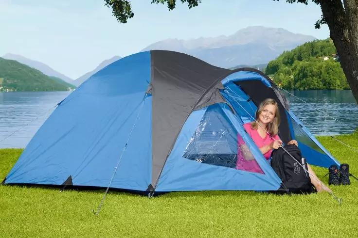 Frau sitzt am Eingang eines blauen Zeltes auf Gras an einem See, Berge im Hintergrund, Baum rechts, Rucksack und Fernglas neben dem Zelt