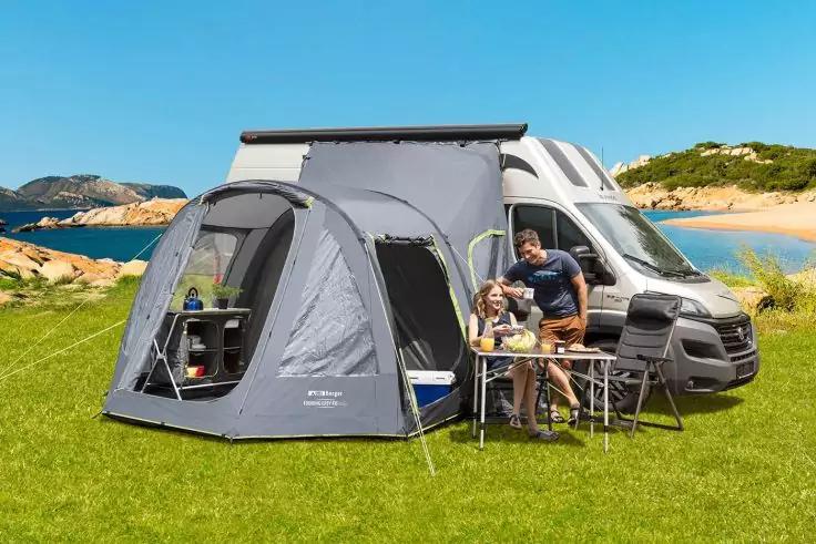 Campingbus mit grauem Vorzelt auf Grasfläche am Wasser; Mann und Kind sitzen an Campingtisch; blauer Himmel und felsige Hügel im Hintergrund