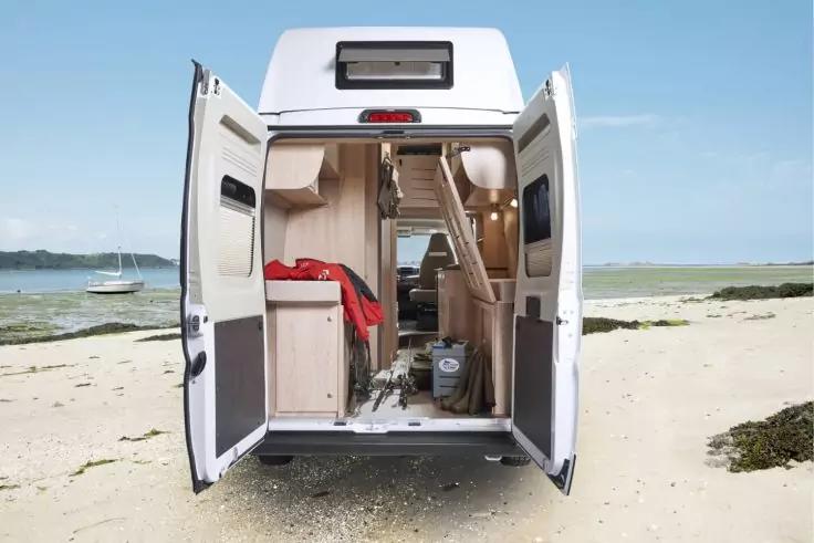 Offenes Heck eines Wohnmobils auf einem Sandstrand, geöffnete Hecktüren, Holzschränke im Innenraum, rotes Tuch, Leiter und das Meer mit einem Segelboot im Hintergrund