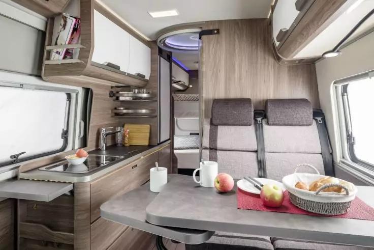 Innenraum eines Wohnmobils mit Küchenzeile (Spüle, Herd, Oberschränke), Tisch mit zwei Bechern, Äpfeln und Brotkorb, zwei gepolsterte Sitze, Fenster und Schlafbereich im Hintergrund