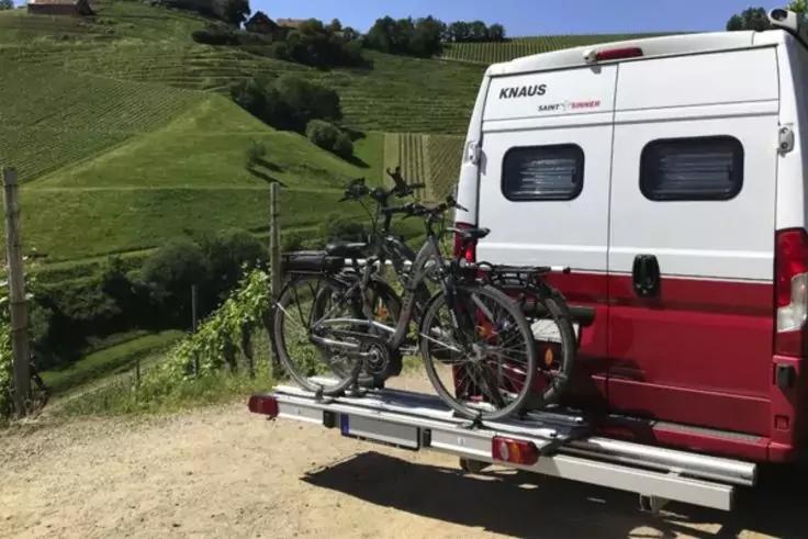 Heckansicht eines wei&szlig;-roten Wohnmobils mit zwei Fahrr&auml;dern auf einem Hecktr&auml;ger, grasbewachsene H&uuml;gel und ein Holzzaun im Hintergrund