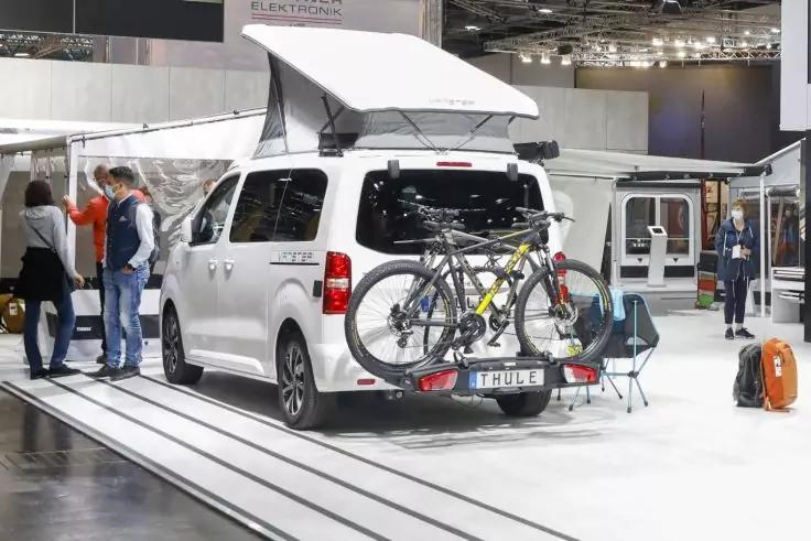 Wei&szlig;er Campervan mit aufgestelltem Aufstelldach, zwei Fahrr&auml;der am Hecktr&auml;ger, Personen links, St&uuml;hle und Gep&auml;ck daneben, weitere Fahrzeuge in Messehalle