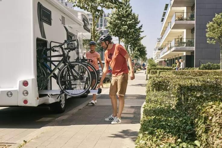 Zwei Personen neben einem wei&szlig;en Wohnmobil; eine Person h&auml;lt ein Fahrrad vom Hecktr&auml;ger; mehrere Fahrr&auml;der am Heck; Gehweg, Hecken und moderne Wohngeb&auml;ude im Hintergrund