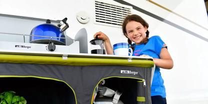 Kind in blauem T-Shirt steht neben einer mobilen Campingk&uuml;che mit gr&uuml;nem Vorhang und h&auml;lt eine Kaffeekanne