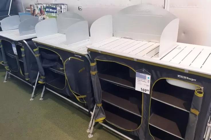 Vier graue, stoffbezogene Einheiten mit gelbem Rand, offenen F&auml;chern und wei&szlig;er Lattenfl&auml;che oben, nebeneinander auf einem wei&szlig;en Podest in einem Gesch&auml;ft; Preisschild sichtbar, gr&uuml;ner Teppichboden