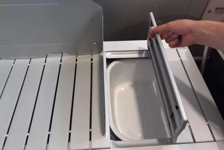 Rechte Hand h&auml;lt eine ge&ouml;ffnete graue Klappe; darunter ein wei&szlig;es rechteckiges Fach; links eine wei&szlig;e Fl&auml;che mit schmalen Schlitzen