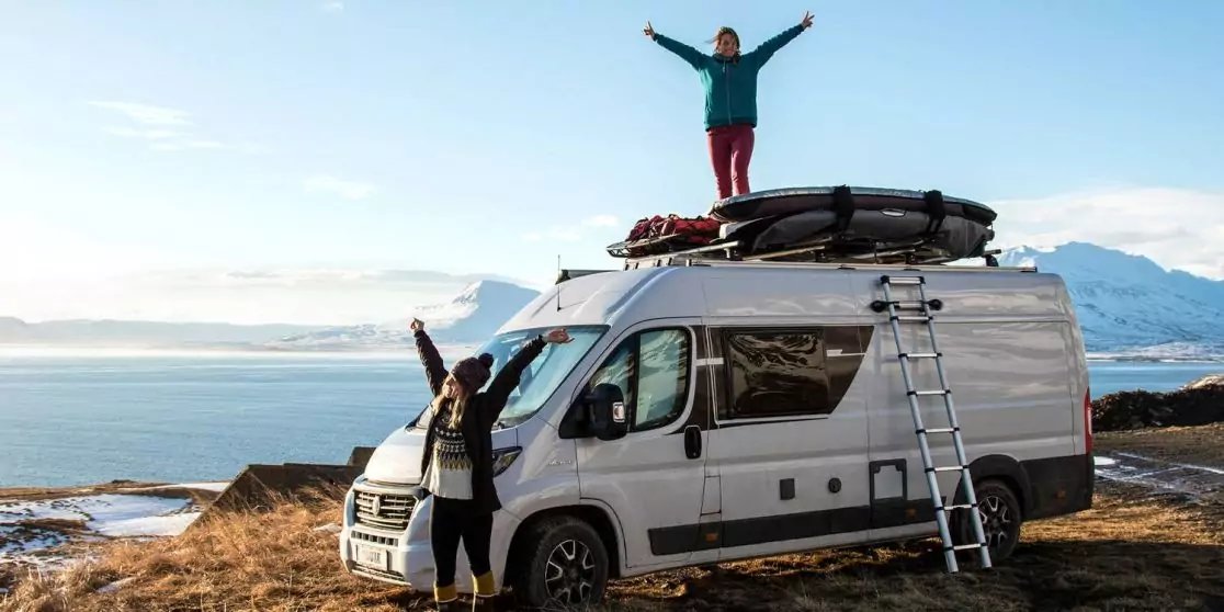 Wei&szlig;er Campervan an der K&uuml;ste vor Meer und schneebedeckten Bergen, Leiter und Ausr&uuml;stung auf dem Dach, zwei Personen, eine steht auf dem Dach mit erhobenen Armen