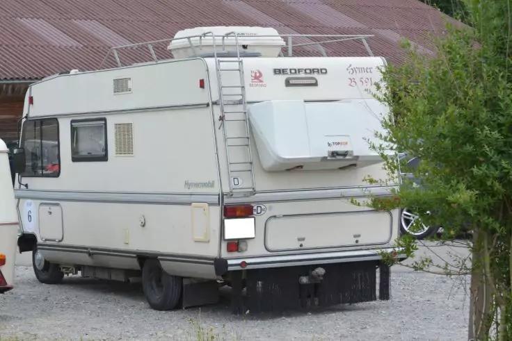 Weißes Wohnmobil, Heck- und Seitenansicht, auf Kiesfläche; Leiter am Heck; Dachbox; Gebäude mit Wellblechdach im Hintergrund; Busch rechts