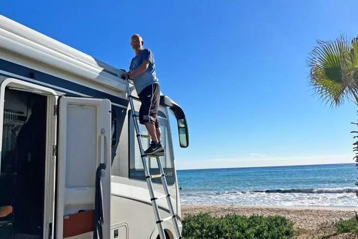 Person auf Leiter bei weißem Wohnmobil am Strand, Meer, blauer Himmel, Palme rechts