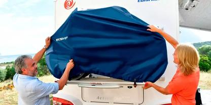 Ein Mann und eine Frau halten einen blauen Bezug &uuml;ber das Heck eines wei&szlig;en Wohnmobils; Wiese und Himmel im Hintergrund