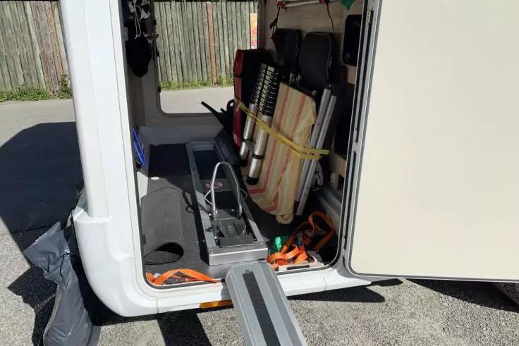 Offenes Seitenfach eines Fahrzeugs mit ausgeklappter Metallrampe bis zum Boden; im Fach liegen zusammengefaltete Ger&auml;te, Gurte und Kabel
