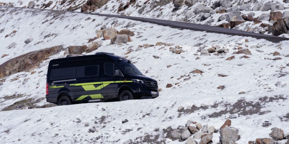 Schwarzer Transporter mit gelb-gr&uuml;nen Markierungen auf schneebedecktem, felsigem Hang; oberhalb verl&auml;uft eine kurvige Stra&szlig;e