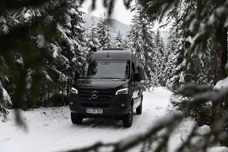 Schwarzer Mercedes-Van auf einer verschneiten Waldstra&szlig;e, umgeben von schneebedeckten Tannen