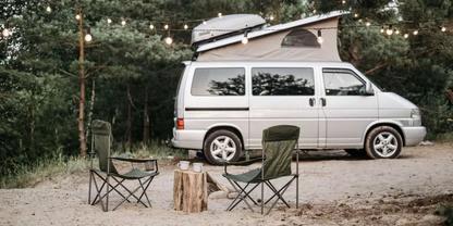 Wei&szlig;er Van mit aufgeklapptem Aufstelldach auf einer Waldlichtung; zwei Klappst&uuml;hle, Baumstumpf als Tisch, Lichterkette &uuml;ber dem Fahrzeug