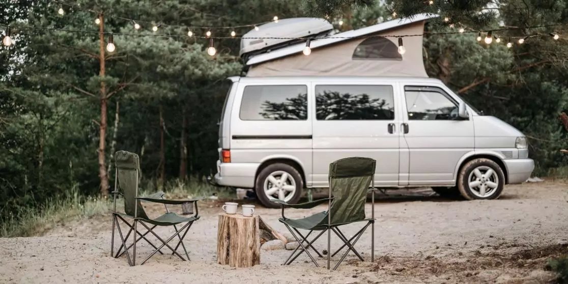 Wei&szlig;er Van mit aufgeklapptem Aufstelldach auf einer Waldlichtung; zwei Klappst&uuml;hle, Baumstumpf als Tisch, Lichterkette &uuml;ber dem Fahrzeug