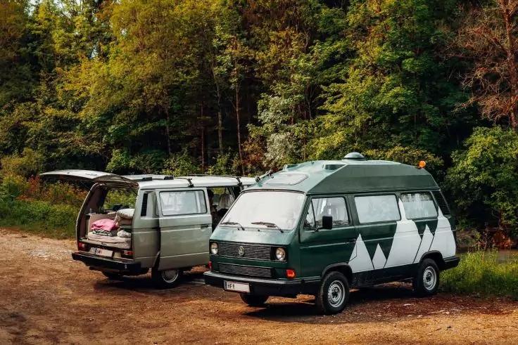 Zwei grüne Campervans auf einem unbefestigten Weg vor einem Wald; eines hat die Heckklappe offen und zeigt Gepäck