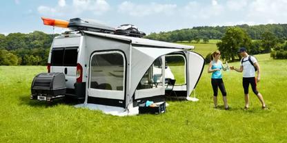 Wei&szlig;er Camper mit grauem Vorzelt, oranger Kajak auf dem Dach, Campingausr&uuml;stung auf dem Gras, zwei Personen in Sportkleidung mit Trinkflaschen auf einer gr&uuml;nen Wiese