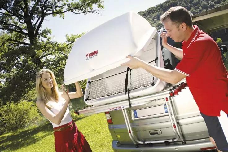 Mann und Frau heben einen ge&ouml;ffneten wei&szlig;en Dachkoffer auf ein silbernes Auto; Gras und B&auml;ume im Hintergrund