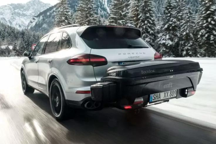 Silbernes Porsche-SUV von hinten auf einer schneebedeckten Stra&szlig;e, schwarzer Hecktr&auml;ger/Koffer an der Anh&auml;ngerkupplung, verschneite Nadelb&auml;ume und Berge im Hintergrund