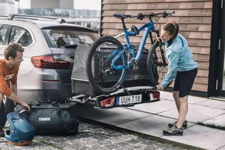 Zwei Personen neben einem silbernen SUV, ein blaues Mountainbike auf einem Hecktr&auml;ger, Fahrradhelm und Tasche auf dem Boden