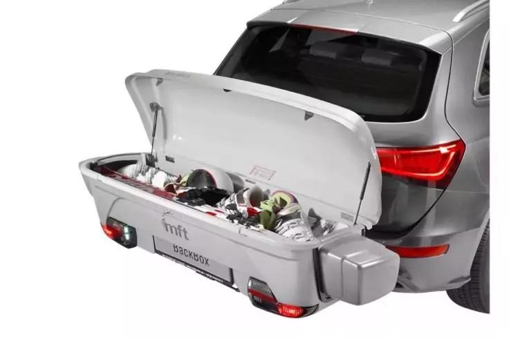 Heck eines silbernen SUV mit offenem, an der Anh&auml;ngerkupplung befestigtem Gep&auml;ckkoffer; im Koffer zwei Fahrradhelme und Taschen sichtbar