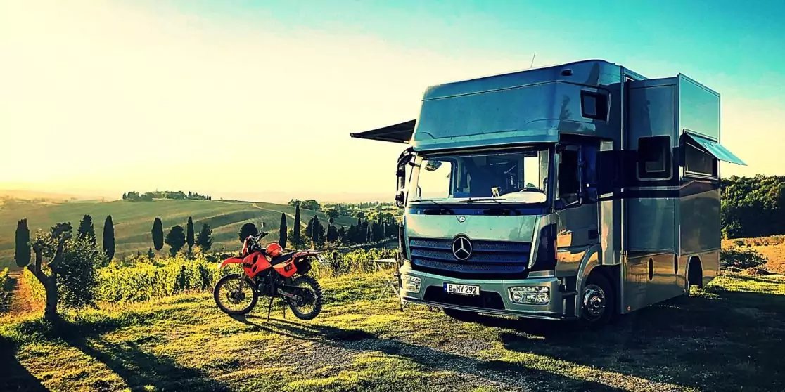 Gro&szlig;er silberner Mercedes-Lkw mit Wohnaufbau und ausgefahrener Seitenmarkise auf einer Wiese, links ein rotes Motorrad, im Hintergrund H&uuml;gel, Reihen von Zypressen und vereinzelte Kakteen, gelb-blauer Himmel
