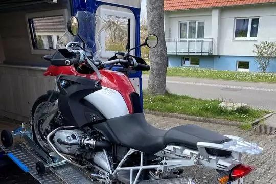 Rotes und wei&szlig;es Motorrad mit schwarzem Sitz auf einer Metallrampe vor einem blauen Fahrzeug; H&auml;user und ein Baum im Hintergrund
