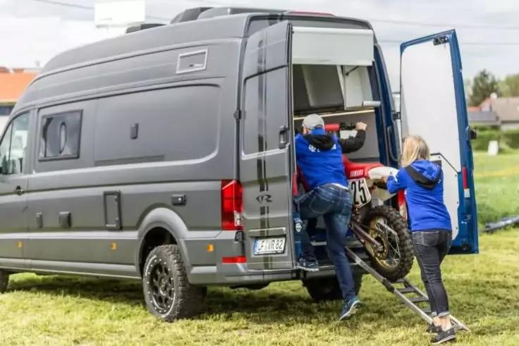 Zwei Personen in blauen Jacken laden ein Motorrad &uuml;ber eine Rampe in den offenen Laderaum eines grauen Vans auf einer Wiese