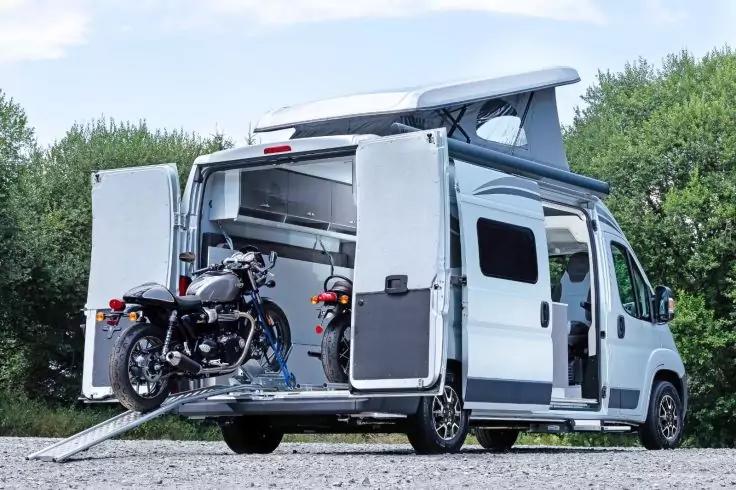 Wei&szlig;er Camper mit offenen Heckt&uuml;ren, aufgeklapptem Aufstelldach und Rampe, auf der ein Motorrad steht, B&auml;ume im Hintergrund