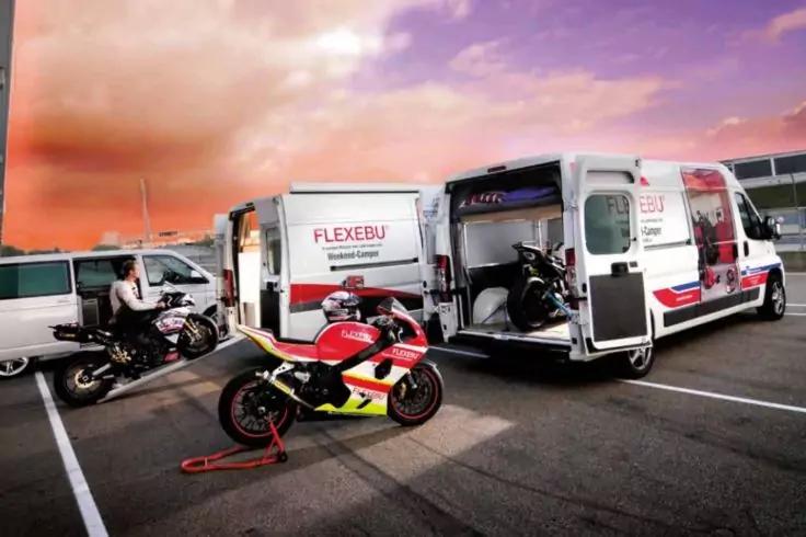 Drei Sportmotorr&auml;der vor zwei wei&szlig;en Transportern mit offenen Heckt&uuml;ren auf einem Parkplatz; eine Person steht neben einem Motorrad; r&ouml;tlich-violetter Himmel im Hintergrund