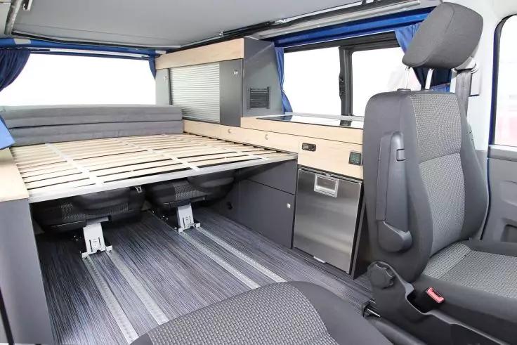 Fahrzeuginnenraum mit zwei vorderen Sitzen, aufgeklappter Lattenrost-Plattform, Holzschr&auml;nken und Schubladen, Fenstern mit blauen Vorh&auml;ngen und grauem gestreiftem Bodenbelag