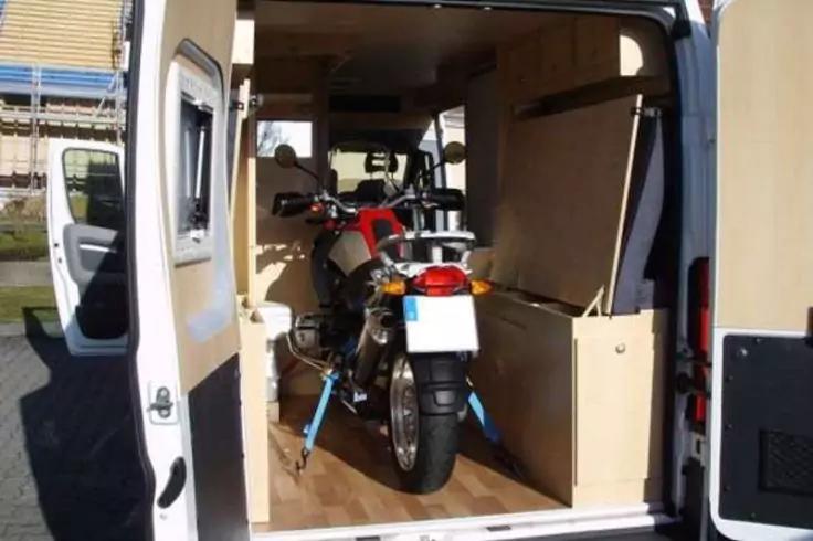 Innenraum eines wei&szlig;en Lieferwagens mit offenen Heckt&uuml;ren, ein rotes Motorrad auf Holzfu&szlig;boden, mit blauen Spanngurten gesichert, seitliche Holzverkleidung und offene Fahrert&uuml;r sichtbar