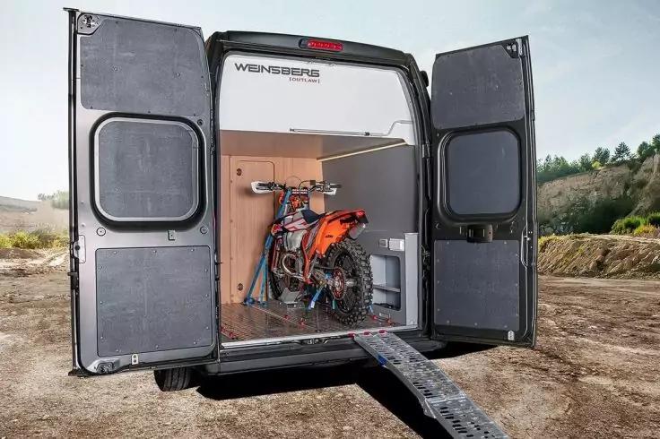 Offenes Heck eines Transporters mit ge&ouml;ffneten Heckt&uuml;ren; im Ladebereich ein oranges Gel&auml;ndemotorrad auf einer Metallrampe, mit Zurrgurten gesichert