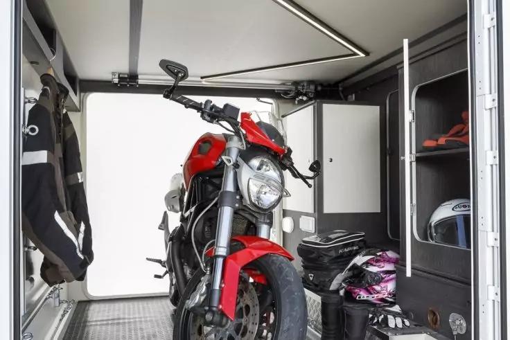 Rotes Motorrad in einem hellen, abgeschlossenen Laderaum mit offener T&uuml;r; rechts Regale mit Helmen und Taschen; links Jacken an der Wand