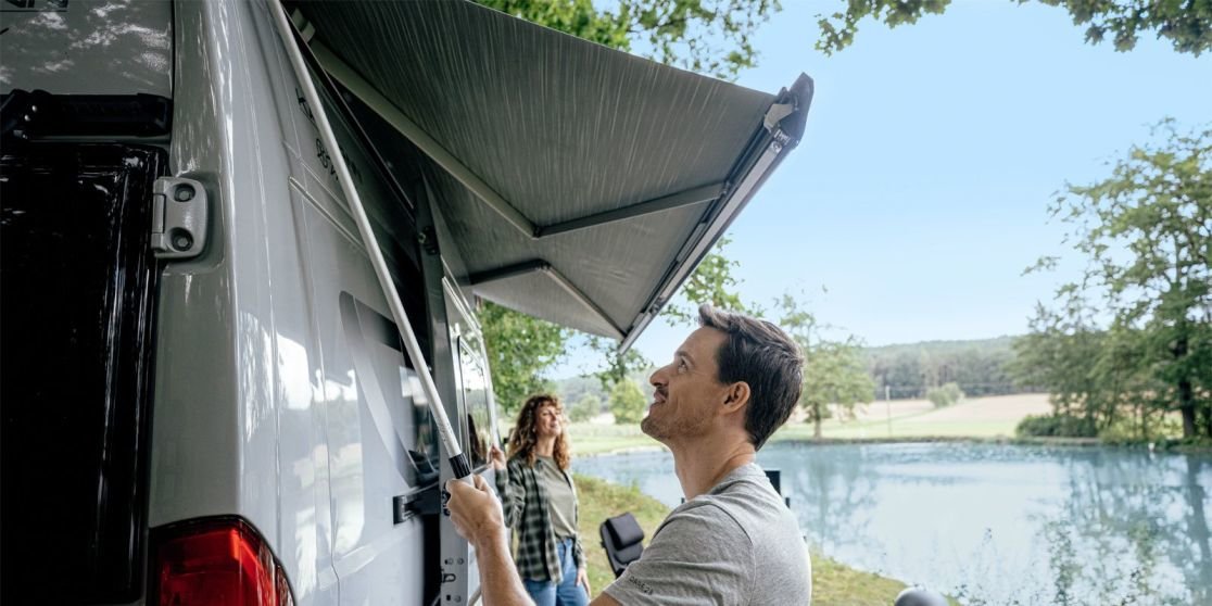 Mann f&auml;hrt die Markise an der Seite eines wei&szlig;en Vans aus, Frau steht daneben, See und B&auml;ume im Hintergrund