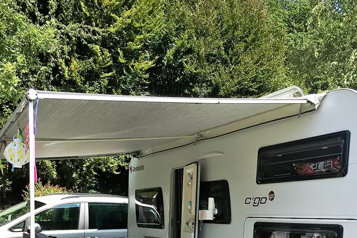 Wei&szlig;er Wohnwagen mit ausgefahrener Markise, links teilweise ein geparktes Auto, im Hintergrund B&auml;ume