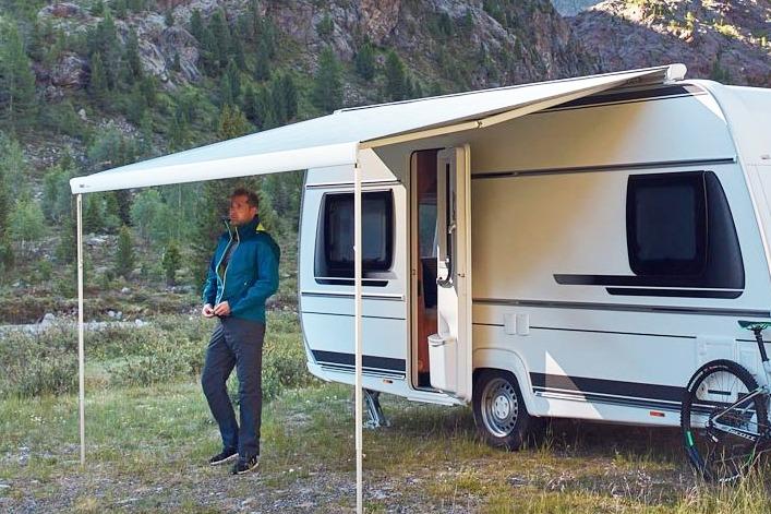 Eine Person in blauer Jacke lehnt am wei&szlig;en Wohnwagen unter ausgefahrener Markise; T&uuml;r ge&ouml;ffnet, Fahrrad am Heck; Gras, B&auml;ume und felsige Bergwand im Hintergrund
